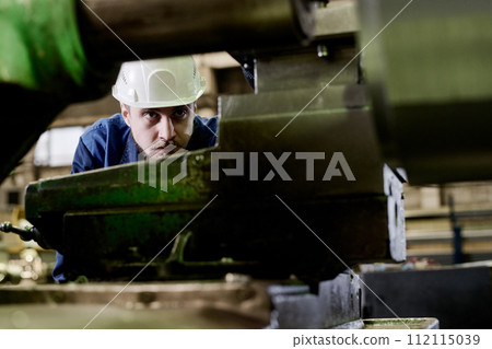 Selective focus shot of serious young man wearing hardhat examining industrial machine Selective focus shot of serious young man wearing hardhat examining industrial machine 112115039