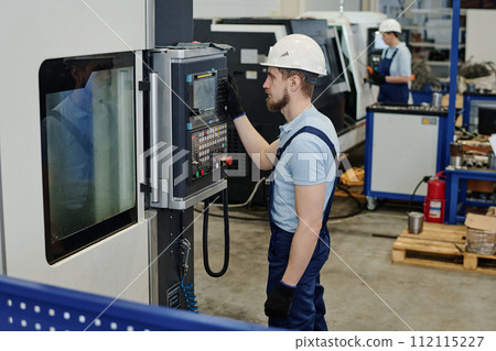 Young male professional turner wearing hardhat operating modern CNC mchine in factory Young male professional turner wearing hardhat operating modern CNC mchine in factory 112115227