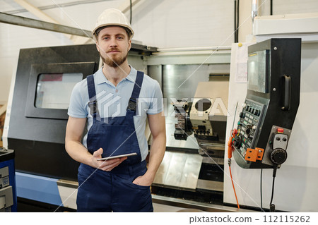 Medium portrait of young bearded factory worker holding digital tablet standing against CNC machine looking at camera Medium portrait of young bearded factory worker holding digital tablet standing against CNC machine looking at camera 112115262