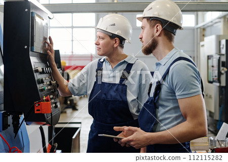 Side view medium shot of male and female engineers wearing hardhats working in factory using CNC machine 112115282