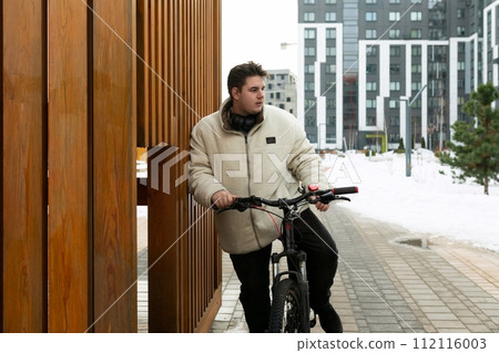 A young European man with a winter jacket walks around the city with a bicycle A young European man with a winter jacket walks around the city with a bicycle 112116003