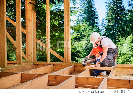 Father with toddler son building wooden frame house near the forest. Man teaching his son how to cut board using electric saw on construction site, wearing overalls. Carpentry and family concept. 112118965