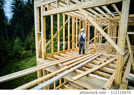 Laborer constructing wooden frame house near forest. Man treating woods, applying fire retardant using sprayer, while dressed in protective suit, helmet. Concept of modern eco-friendly construction. Laborer constructing wooden frame house near forest. Man treating woods, applying fire retardant using sprayer, while dressed in protective suit, helmet. Concept of modern eco-friendly construction. 112118970