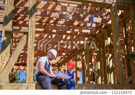 Father with toddler son building wooden frame house. Guys having fun on edge of balcony, examining construction plan, wearing helmets and overalls on sunny day. Carpentry and family concept. Father with toddler son building wooden frame house. Guys having fun on edge of balcony, examining construction plan, wearing helmets and overalls on sunny day. Carpentry and family concept. 112118975