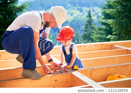 Father with toddler son building wooden frame house. Male builder teaching his son how to measure angle on construction site, wearing helmets and overalls on sunny day. Carpentry and family concept. 112118976