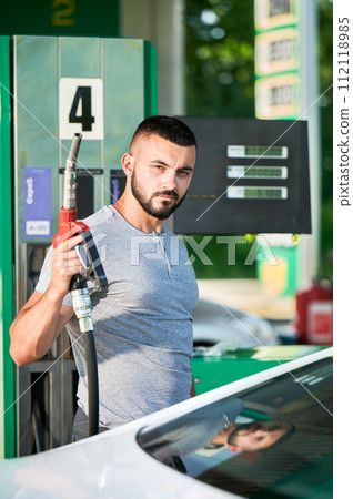 Skilled driver refueling his automobile for long road. Portrait of male adult with pump nozzle refueling car tank. Man holding gasoline pump next to car on background gas column. Skilled driver refueling his automobile for long road. Portrait of male adult with pump nozzle refueling car tank. Man holding gasoline pump next to car on background gas column. 112118985