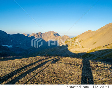 Champery, Switzerland - September 10th 2023: Three shadow hikers enjyoing alpine early morning view. Champery, Switzerland - September 10th 2023: Three shadow hikers enjyoing alpine early morning view. 112120961