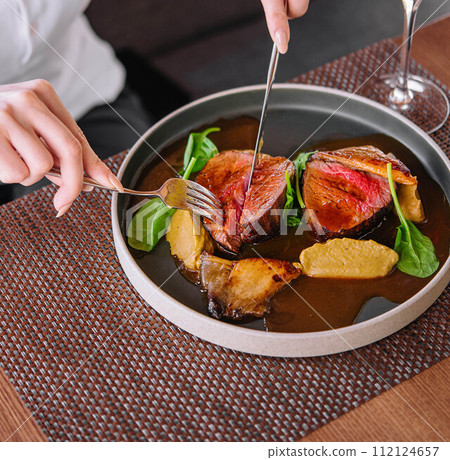 female hands holding a fork and knife and cuts roast duck with wine 112124657