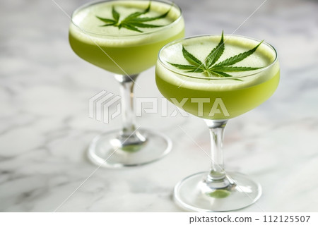 Two elegant cocktails with frothy tops, each adorned with a cannabis leaf on a marble surface 112125507