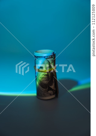 One hundred dollar bills rolled into a tube and wrapped with an elastic band under hard blue light 112125809
