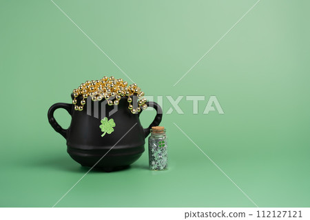 Black cauldron pot with a glitter clover and gold beads and a phial with silver starts on green background. Design element detail for magic fantasy photo composits or St Patrick's 112127121