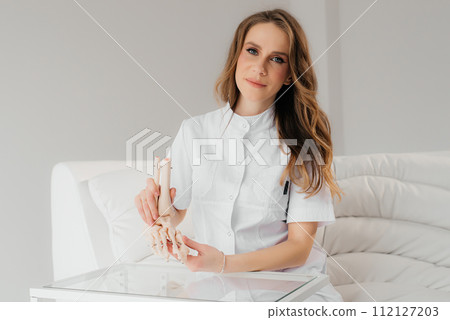 A doctor studies a womans leg structure while holding model of skeleton on a white table indoors. Models of skeletons help doctors study the structure of the human body 112127203