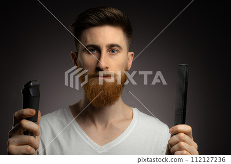 Man shaving whiskers hair by himself, self haircut at home in front of mirror in the bath. Male with long haircut using shaving machine trimmer. 112127236