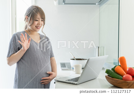 Pregnant woman having a video call/video call on a laptop in the kitchen (waving, greeting, cooking class) Pregnant woman having a video call/video call on a laptop in the kitchen (waving, greeting, cooking class) 112127317
