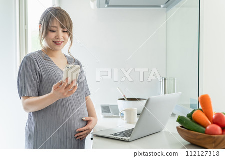 Pregnant woman using computer and smartphone while cooking in the kitchen Pregnant woman using computer and smartphone while cooking in the kitchen 112127318