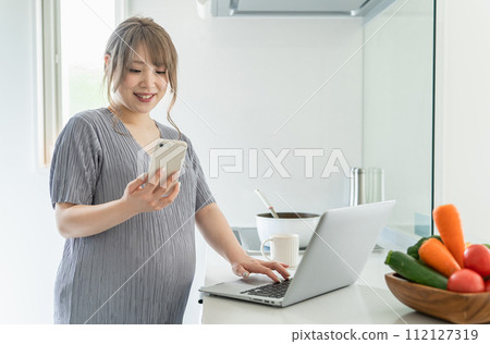 Pregnant woman using computer and smartphone while cooking in the kitchen Pregnant woman using computer and smartphone while cooking in the kitchen 112127319