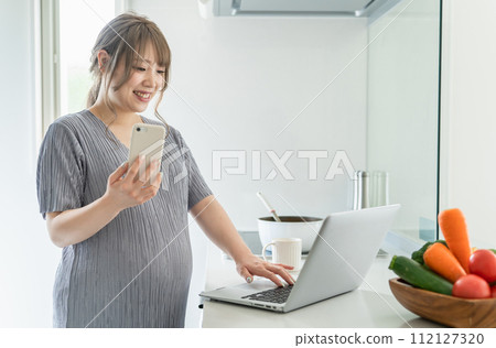 Pregnant woman using computer and smartphone while cooking in the kitchen 112127320