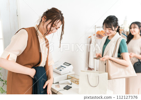 一家服飾店的女店員在收銀台折疊牛仔布,女顧客拿著智慧型手機排隊 一家服飾店的女店員在收銀台折疊牛仔布,女顧客拿著智慧型手機排隊 112127376
