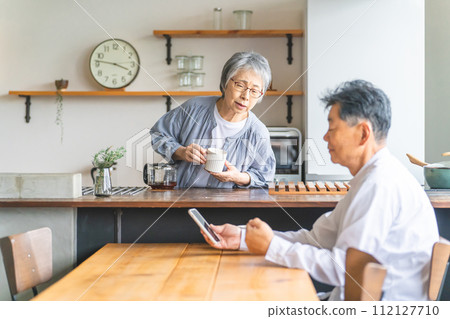 A senior woman/wife looks worriedly at her husband who uses a smartphone in the dining room A senior woman/wife looks worriedly at her husband who uses a smartphone in the dining room 112127710