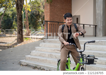 Asian businessman in a suit is riding a bicycle on the city streets for his morning commute to work. Eco transportation concept, sustainable lifestyle concept Asian businessman in a suit is riding a bicycle on the city streets for his morning commute to work. Eco transportation concept, sustainable lifestyle concept 112128705