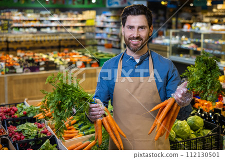A skilled mature male employee in a supermarket stands proudly in the produce section, with a variety of fresh vegetables surrounding him, signaling quality and freshness. A skilled mature male employee in a supermarket stands proudly in the produce section, with a variety of fresh vegetables surrounding him, signaling quality and freshness. 112130501
