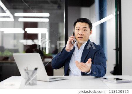 Adult entrepreneur having phone conversation while sitting bu equipped table with computer in corporate building. Calm startup leader discussing project details with perspective investor on distance. 112130525