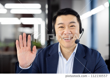 First person view of mature eastern man wearing headset with microphone and making greeting gesture to camera. Smiling formal male talking on video call with colleagues and waving hello indoors. 112130557