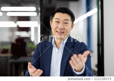 Adult office administrator in business suit looking at camera with friendly smile and gesturing with hands. Hosting company receptionist welcoming partners of boss and asking to join in boardroom. 112130573