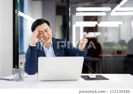 Frustrated chinese boss of company touching forehead with hand while squinting in pain at workplace. Stressed formal male asking employee for bringing medicine for migraine attack relieving. 112130580