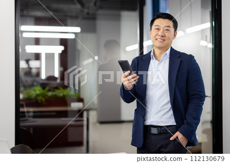 Portrait of successful young Asian man in suit standing in office, holding hand in pocket and mobile phone in other hand, looking confidently and smiling at camera. Portrait of successful young Asian man in suit standing in office, holding hand in pocket and mobile phone in other hand, looking confidently and smiling at camera. 112130679