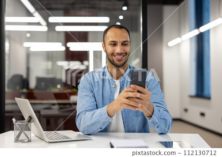 A young male professional in a casual blue denim shirt pauses work to look at his phone. The modern office setting has a relaxed yet productive ambiance. 112130712