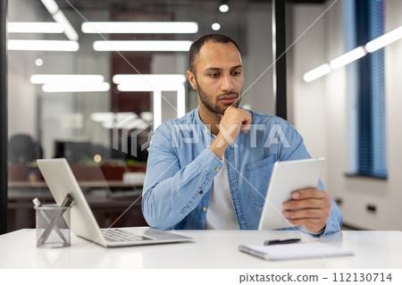 A professional male entrepreneur in a blue shirt is absorbed in reading financial reports on a digital tablet at his workspace, illustrating the concept of business analysis, digital workflow, and 112130714