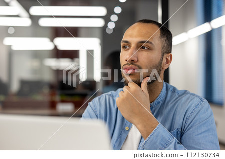 A contemplative young professional in a well-lit office environment, caught in a moment of thoughtfulness while gazing away from his laptop screen. His expression conveys concentration and deep 112130734