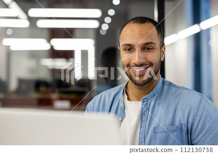 A young, professional man wearing a denim shirt smiles confidently at the camera from his workstation in a contemporary office environment, illuminated by natural and artificial light, embodying A young, professional man wearing a denim shirt smiles confidently at the camera from his workstation in a contemporary office environment, illuminated by natural and artificial light, embodying 112130738