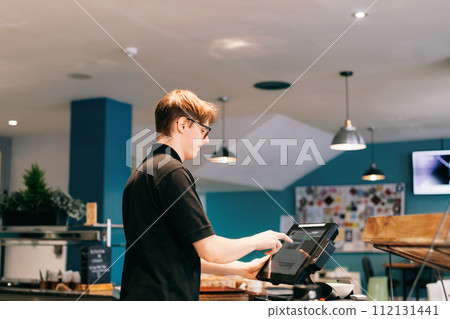 Young waiter serving customer at cash point in cafe. Man working with POS terminal. Cashier, barista checking for payment receipt. Hospitality, server and preparing a slip at the till in coffee shop Young waiter serving customer at cash point in cafe. Man working with POS terminal. Cashier, barista checking for payment receipt. Hospitality, server and preparing a slip at the till in coffee shop 112131441