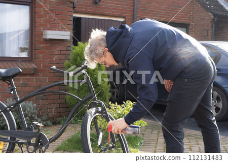 Young man in casual clothes cleaning rust and oiling his city cruiser bicycle chain and gear with spray before riding. Male doing DIY maintenance of his bicycle at home. Selective focus. 112131483