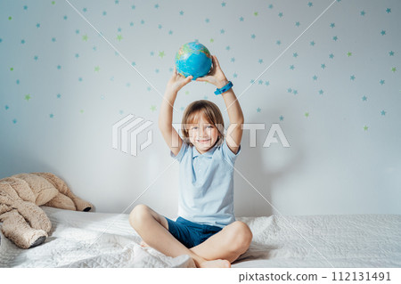 Smiling caucasian boy holding an earth globe model over his head sitting on the bed with stars background on the white wall. Save the planet, Earth Day. Future Global peace and unity concept 112131491