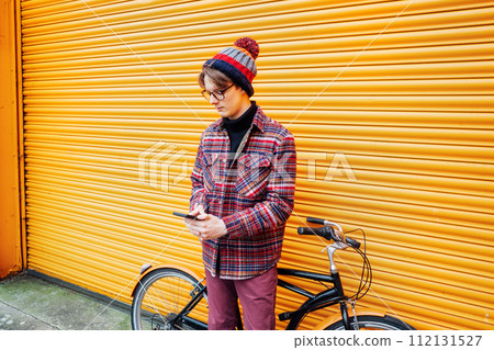 Young hipster man using mobile phone while sitting on city bicycle near orange wall. Student using 5g network,rests after riding. Eco friendly transport for carbon footprint. Sustainable lifestyle 112131527