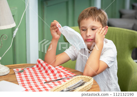 A little boy sits at the table and holds a medical mask in his hand. A little boy sits at the table and holds a medical mask in his hand. 112132190
