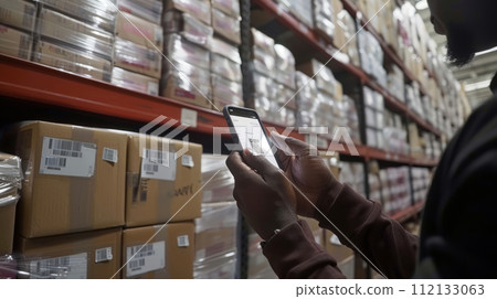 Warehouse Employee Verifies Stock Using a Phone app Amidst Shelves of Boxed Goods. Warehouse Employee Verifies Stock Using a Phone app Amidst Shelves of Boxed Goods. 112133063