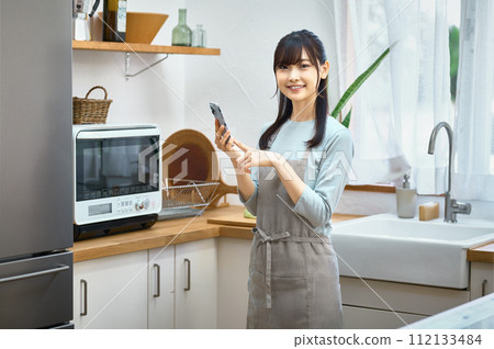 A young housewife wearing an apron looking at a smartphone in the kitchen A young housewife wearing an apron looking at a smartphone in the kitchen 112133484