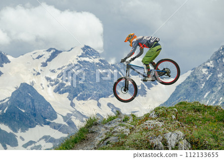 Mountain biker defies gravity, soaring mid-air against backdrop of snow-capped peaks and lush greenery. Ideal for adventure, sports, and nature themes 112133655