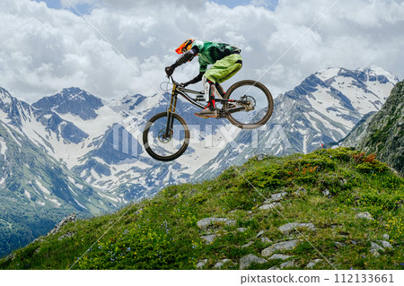 Mountain biker jumped, soaring mid-air against backdrop of snow-capped peaks and lush greenery. downhill competition 112133661