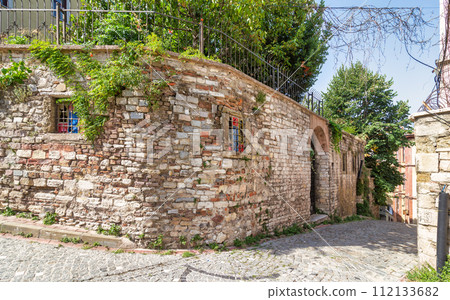 Old abandoned building, with stone wall, wrought iron windows, and climber plants, in cobblestone alley 112133682