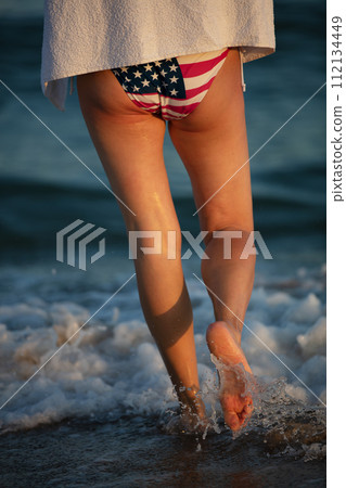 Woman legs in bikini with usa flag go along the sea. America vacation concept on the ocean. Woman legs in bikini with usa flag go along the sea. America vacation concept on the ocean. 112134449