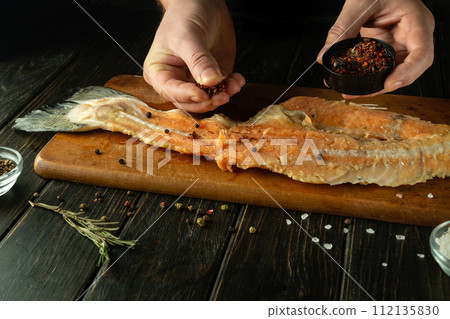 Close-up of a man hands adding aromatic spices to a salmon steak for flavor. Concept of preparing a fish dish on the kitchen table at home Close-up of a man hands adding aromatic spices to a salmon steak for flavor. Concept of preparing a fish dish on the kitchen table at home 112135830