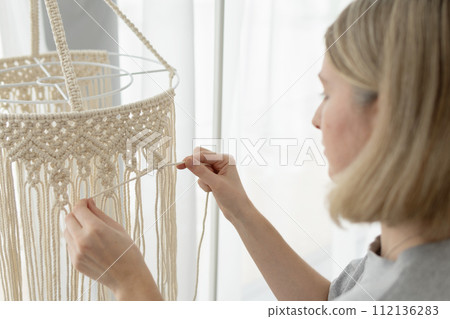 Woman making macrame lampshade using white cord to tie the strings together. Woman knits lampshade for interior using macrame technique Woman making macrame lampshade using white cord to tie the strings together. Woman knits lampshade for interior using macrame technique 112136283