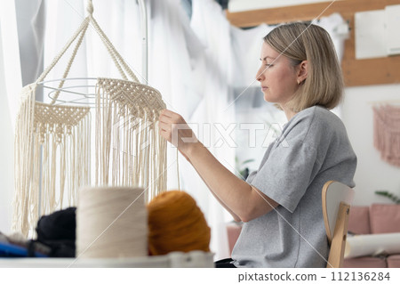 Woman making macrame lampshade using white cord to tie the strings together. Woman knits lampshade for interior using macrame technique Woman making macrame lampshade using white cord to tie the strings together. Woman knits lampshade for interior using macrame technique 112136284