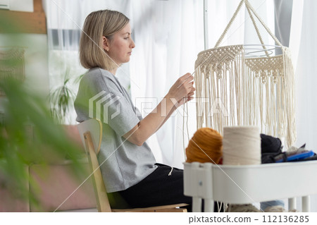 Middle-aged woman knits boho lampshade for interior using macrame technique. Middle-aged woman knits boho lampshade for interior using macrame technique. 112136285