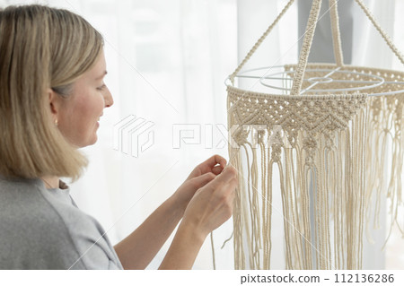 A slender beautiful middle-aged woman knits products for the interior using macrame technique A slender beautiful middle-aged woman knits products for the interior using macrame technique 112136286
